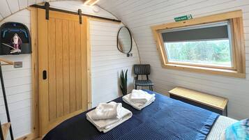A compact room with a bed, a chair, a small table, a wardrobe, a mirror, and a window with a view of trees.