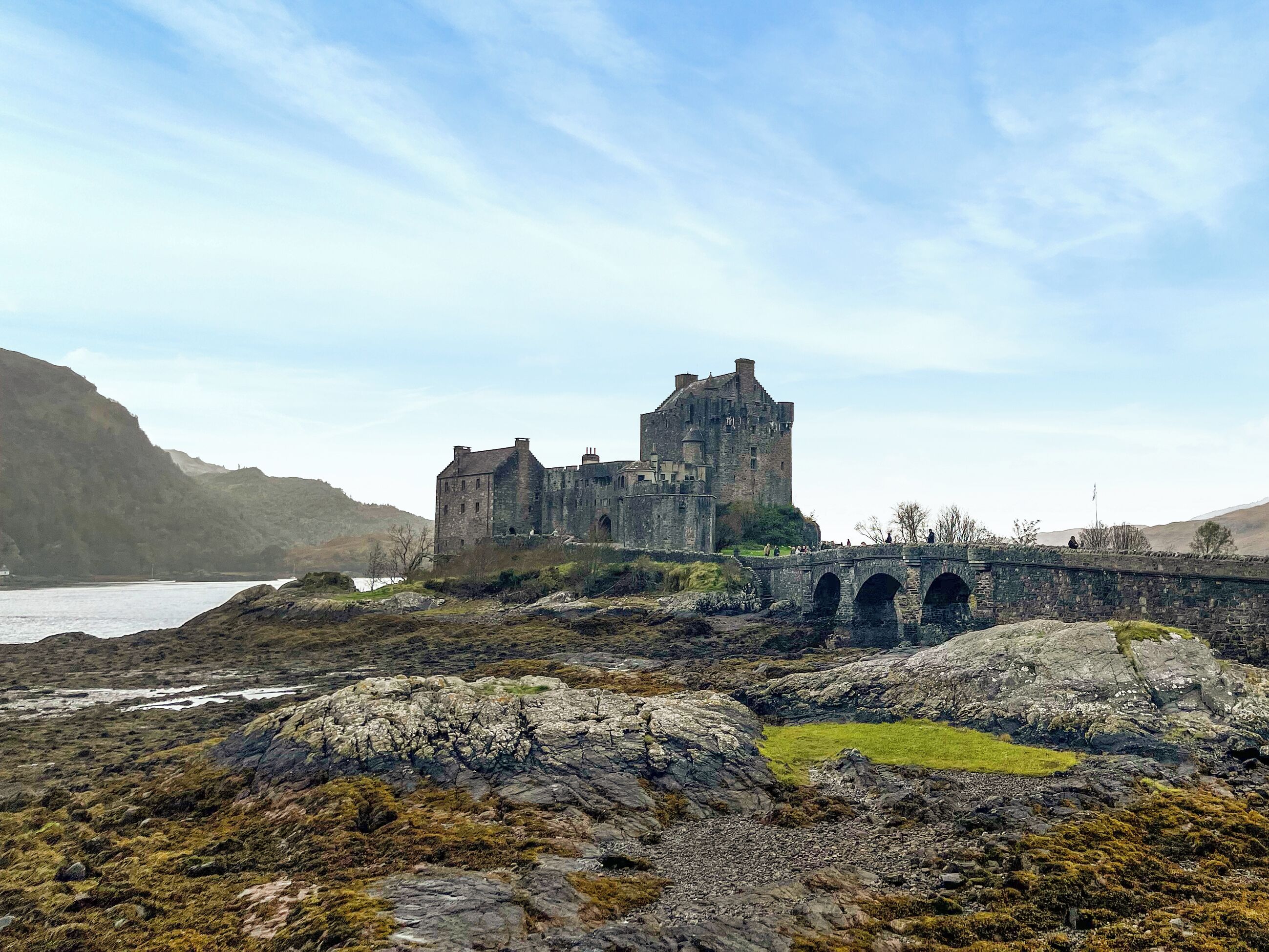 A historic stone castle with a bridge leading to it, surrounded by rocky terrain and a body of water.