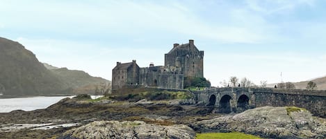A historic stone castle with a bridge leading to it, surrounded by rocky terrain and a body of water.