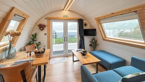Un interior acogedor con una mesa de madera, asientos azules, un televisor y vistas a través de puertas de vidrio.