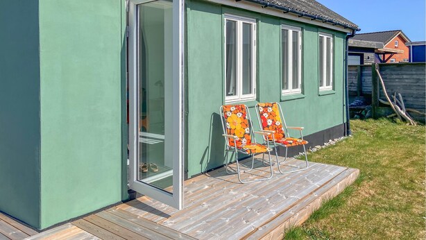 Een groen huis met een houten terras, twee stoelen met bloemenprint bekleding, en een schuifdeur van glas.