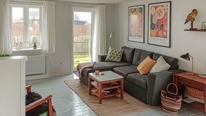 Een gezellige woonkamer met een grijze bank, een klein houten bijzettafeltje en een rode fauteuil. Twee ingelijste foto’s hangen aan de muur, een lamp staat op het tafeltje en er staat een mand op de vloer.