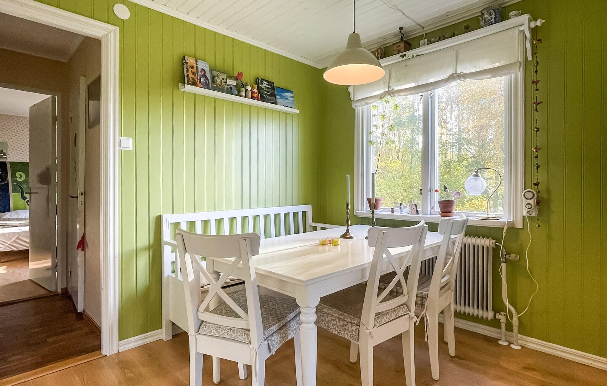A dining area with a white table and chairs, a white bench, a window with a view of greenery, and a shelf with books and decorative items.