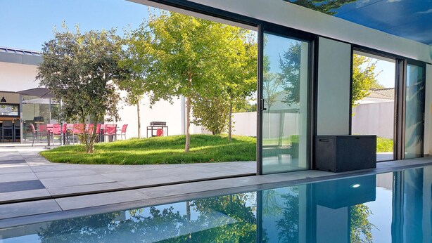 A modern outdoor pool area with clear glass walls reflecting trees and a patio with red chairs. - Villa piscine intérieure (Royan)