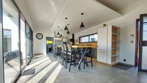 A modern kitchen with a dining area, featuring a large window, pendant lights, and a clock on the wall. - Villa piscine intérieure (Royan)