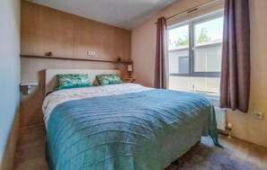 Een slaapkamer met een bed, nachtkastje, lamp, raam met gordijnen en een wandplank.