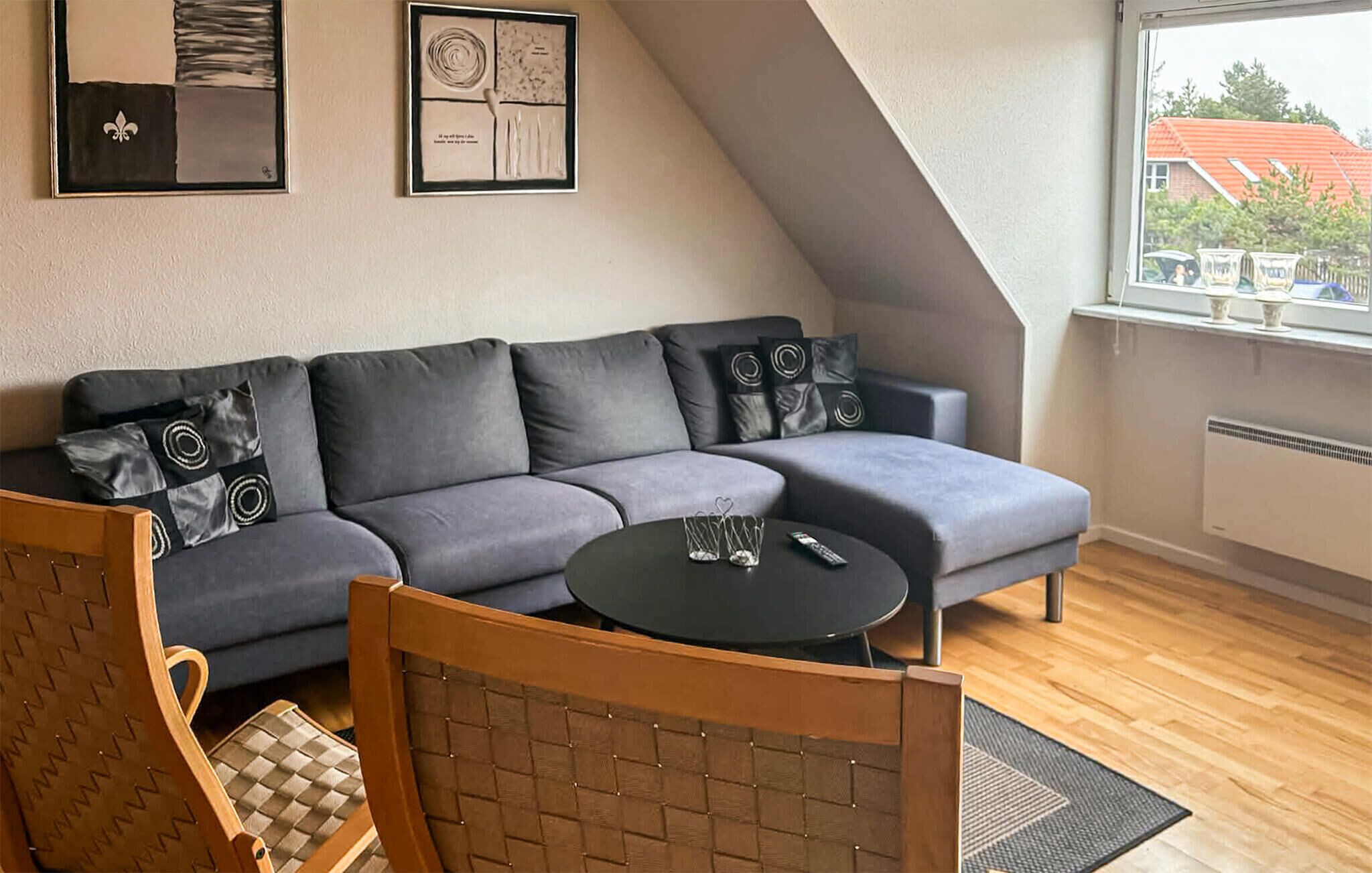 Un salon moderne avec un canapé gris, une chaise en bois, une petite table ronde et une fenêtre donnant sur un bâtiment au toit rouge.