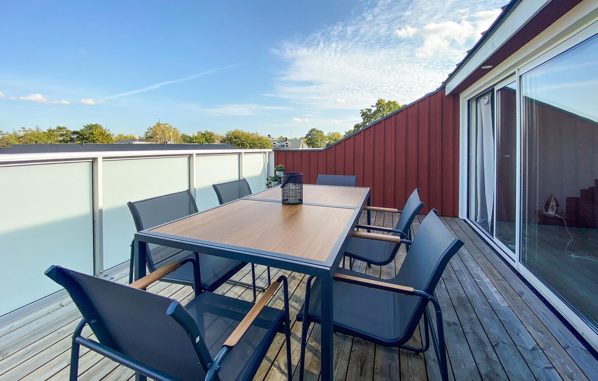 Une terrasse avec une table et des chaises en bois, un pot de fleurs et une porte vitrée donnant sur un espace abrité.