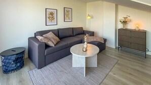 A modern living room with a grey sofa, a round coffee table, and a patterned rug. There are two framed artworks on the wall, a lamp, and a cabinet with a vase of flowers.