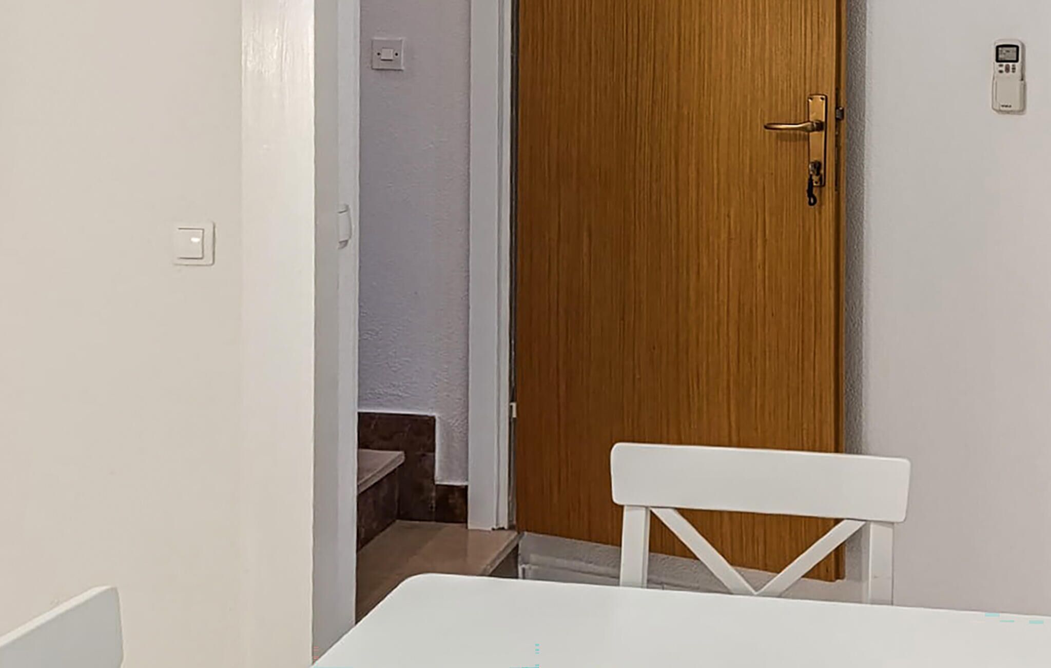 Un couloir étroit mène à une porte en bois dotée d’une poignée et d’une serrure. Une chaise blanche est placée au premier plan.
