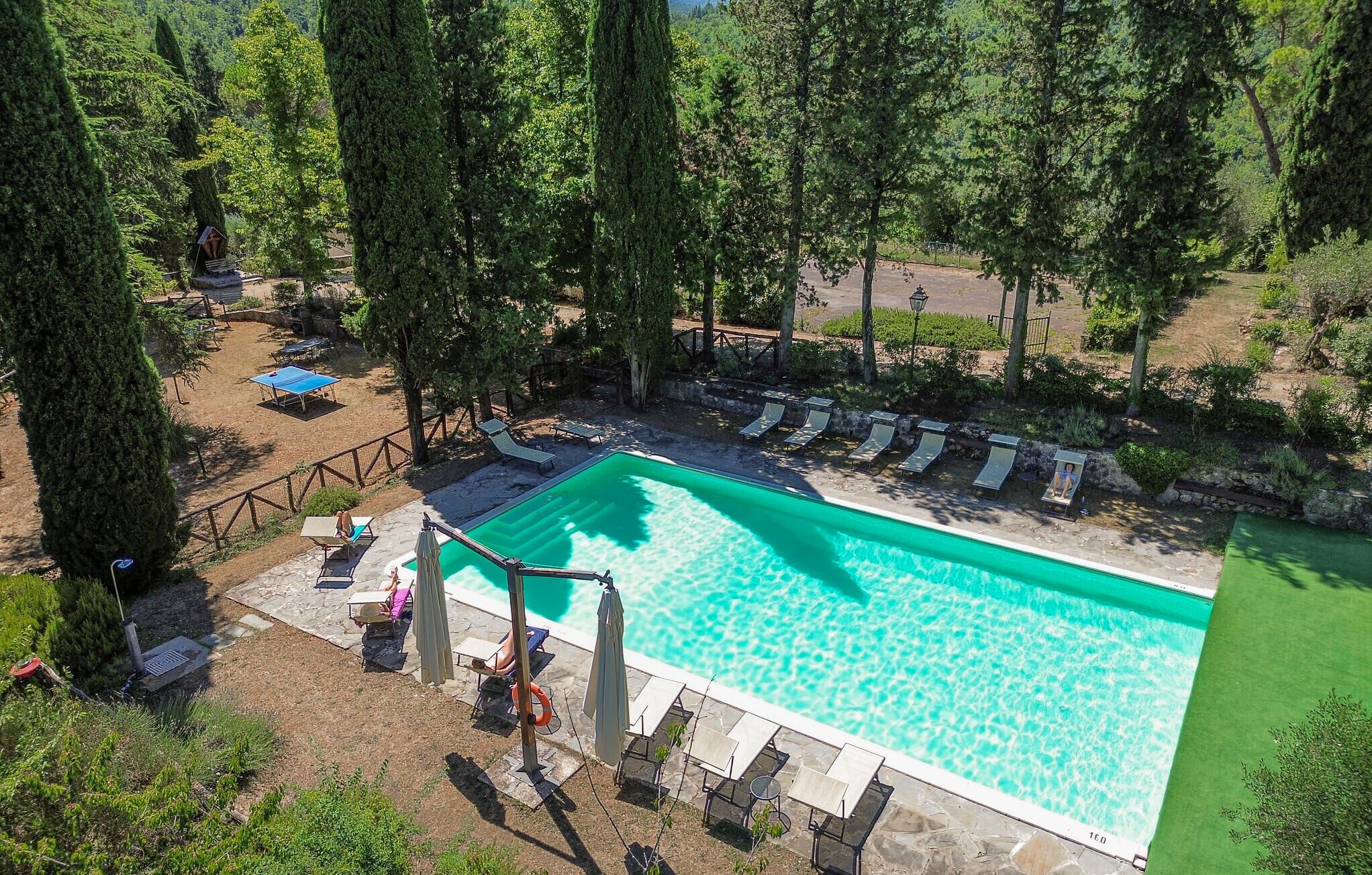 A rectangular swimming pool surrounded by lounge chairs and umbrellas, with a stone deck and a wooden fence.