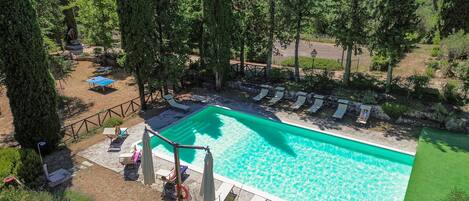 A rectangular swimming pool surrounded by lounge chairs and umbrellas, with a stone deck and a wooden fence.