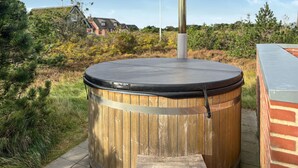Un bain à remous en bois, doté d’un couvercle noir, d’une cheminée en métal et d’une structure en briques en arrière-plan.