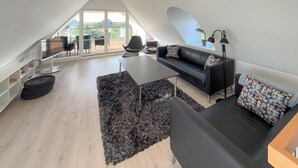 Un salon moderne de style industriel, avec un canapé en cuir noir, une table  à manger en verre et une grande fenêtre donnant sur l’extérieur.