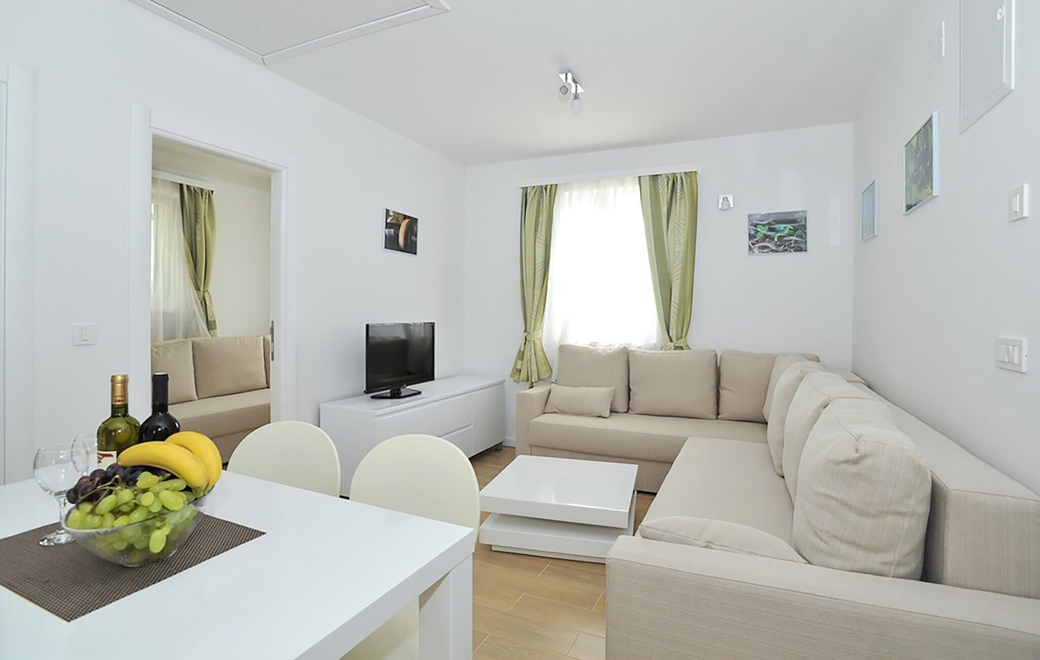 A modern living room with a beige sofa, a white coffee table, and a TV on a white stand. There is a dining table with a bowl of fruit and wine glasses.