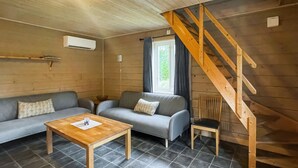 Et hyggeligt værelse i sommerhusstil med en trætrappe, en grå sofa, et træbord og et vindue med gardiner.