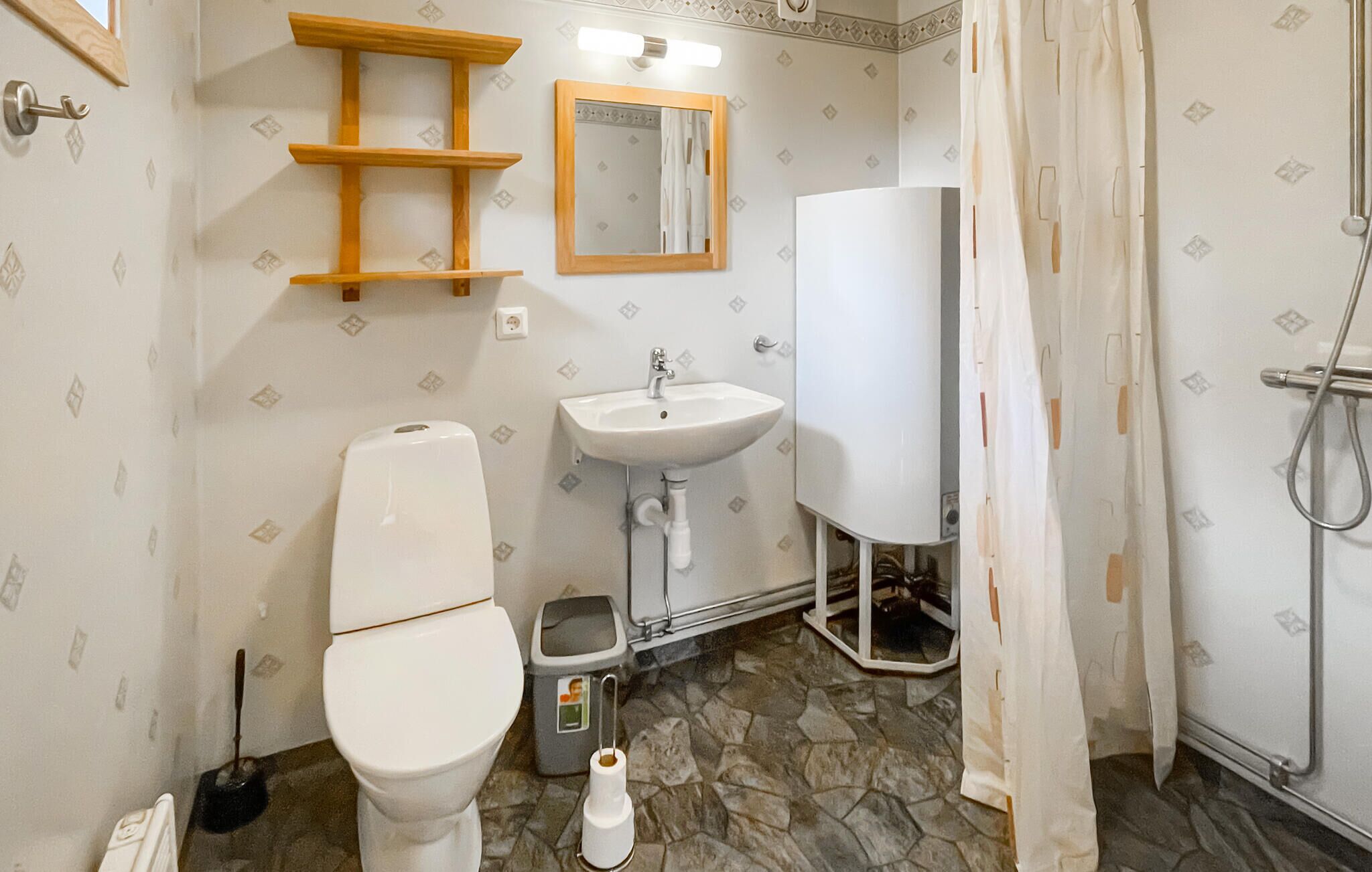 Et badeværelse med toilet, vask og bruser. Der er en træhylde over toilettet og et spejl over vasken. Gulvet er fliset.