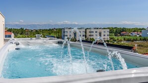A hot tub with multiple jets, surrounded by a pool, with a view of buildings and mountains in the background. - Amazing home in Privlaka with sauna (Privlaka)