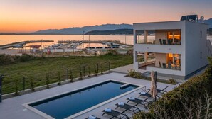 Un bâtiment moderne à deux étages doté d’une piscine et d’un espace extérieur aménagé, offrant une vue sur une marina et un plan d’eau.