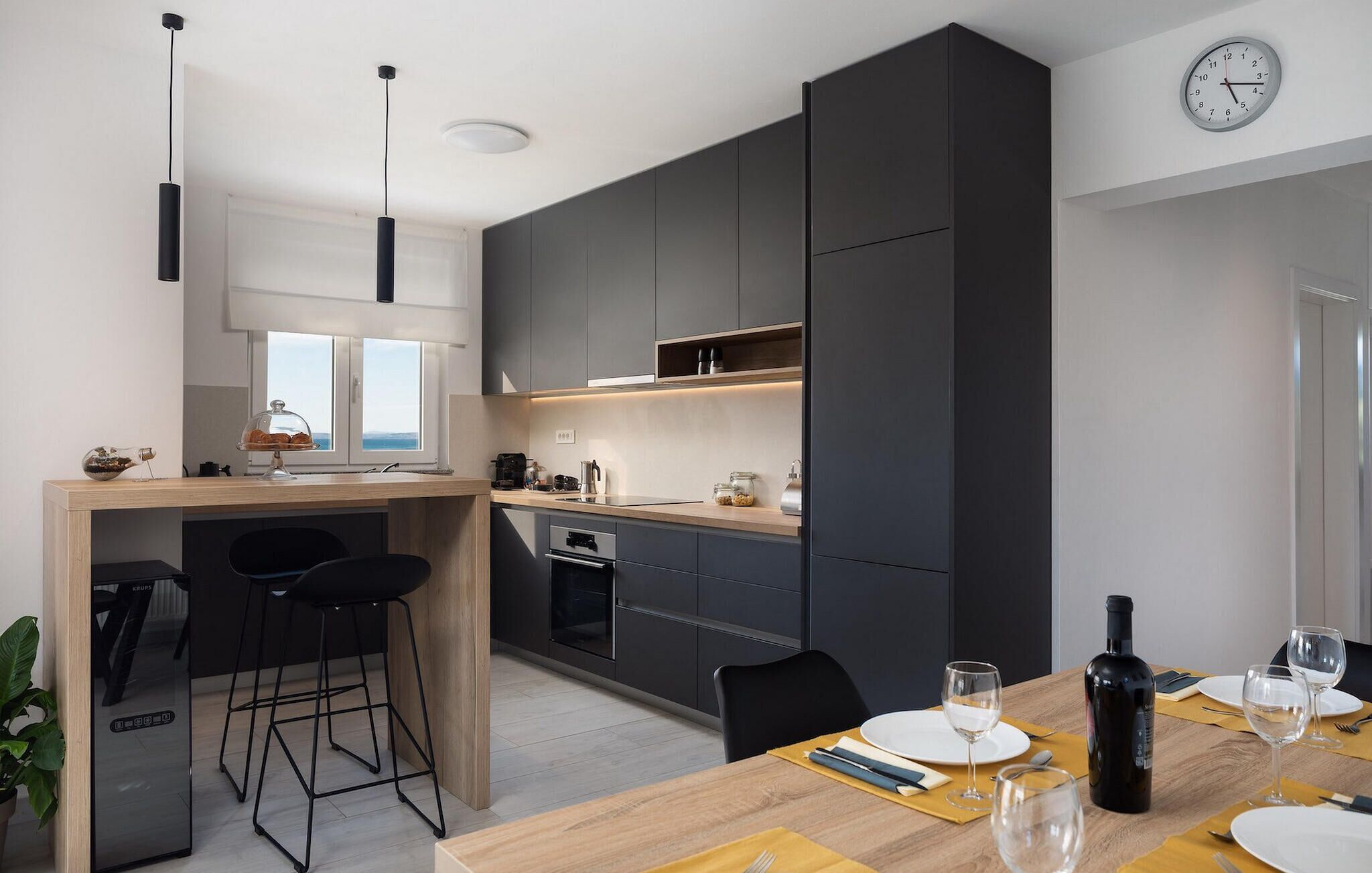 A modern kitchen with dark cabinets, a wooden dining table, and a view of the sea through the window.