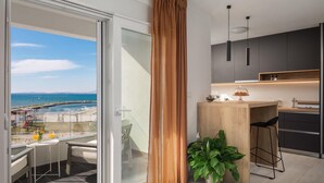 Une cuisine moderne avec un îlot central en bois, un coin repas avec une table et des chaises, et une vue sur la mer grâce à une porte-fenêtre coulissante.
