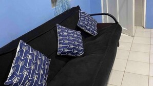 Une pièce avec un canapé noir, deux coussins à motifs, une fenêtre avec un rideau bleu et une porte donnant sur une autre pièce.