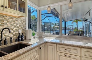 A kitchen with a granite countertop, a sink, and a backsplash. There are cabinets with glass doors and a hanging light fixture. - No Bad Days - Boat Dock, Heated Pool & Hot tub, Beach Gear, 5 Mins to AMI (Bradenton)