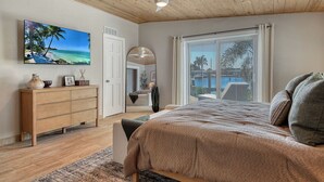A bedroom with a large bed, a ceiling fan, a wooden dresser, a TV displaying a beach scene, and a view of the outdoors through a large window. - No Bad Days - Boat Dock, Heated Pool & Hot tub, Beach Gear, 5 Mins to AMI (Bradenton)