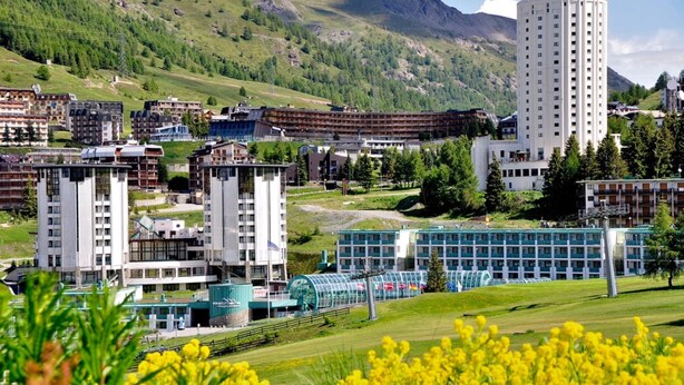 A hotel with multiple buildings, a prominent cylindrical tower, and a green landscape with hills in the background. - Studio Valleverde 11D Ski In Ski Out (Sestriere)