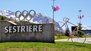 A sign with 'SESTRIERE' and Olympic rings, a bicycle, and flags against a mountain backdrop. - Studio Valleverde 11D Ski In Ski Out (Sestriere)