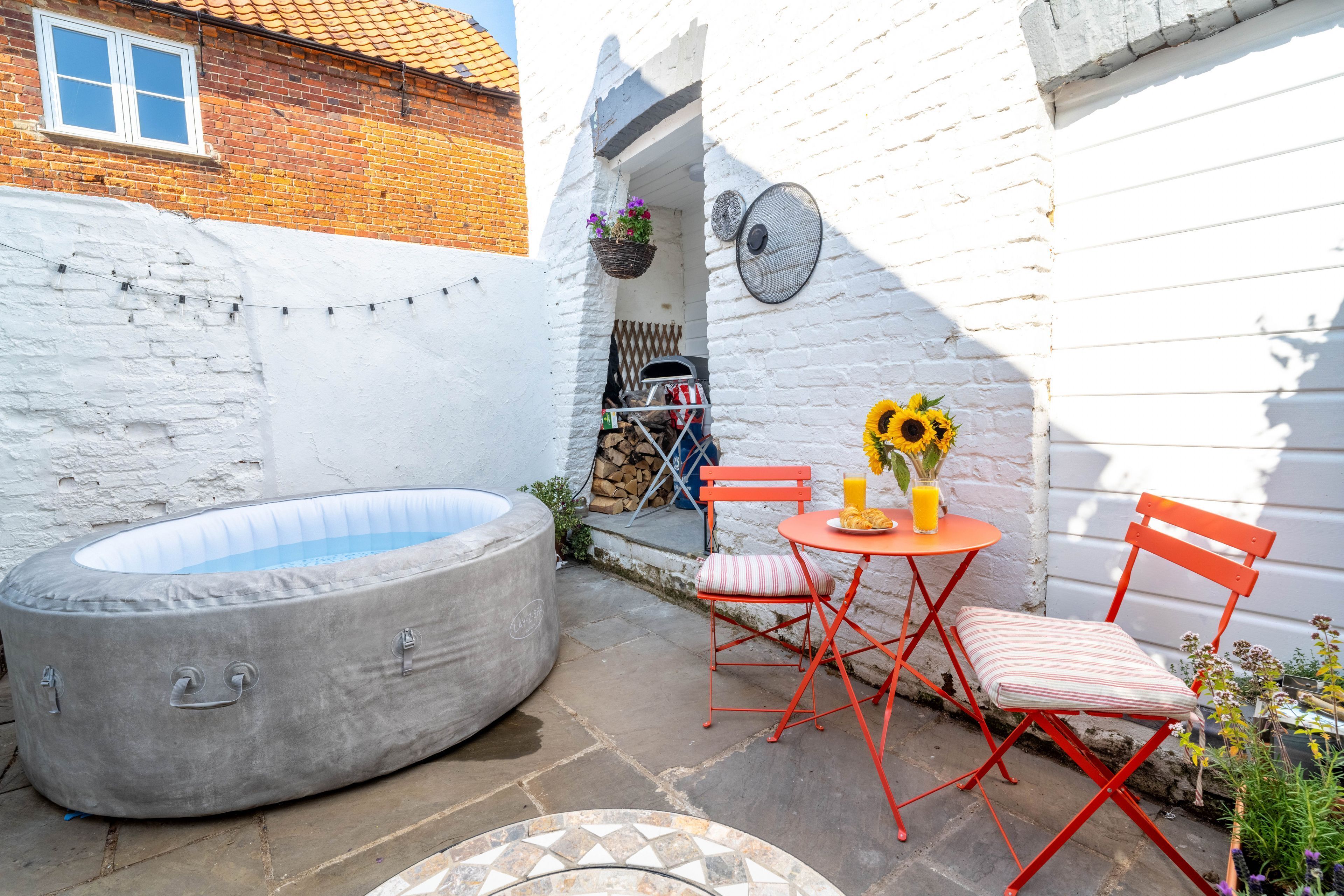 A small outdoor patio with a hot tub, a table with chairs, and a white wall.