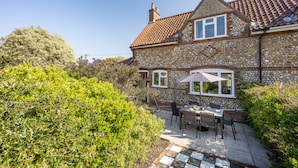 A stone house with a tiled roof, a patio with outdoor furniture, and a garden. - Fairstead Cottage, Holt, Norfolk (Holt)