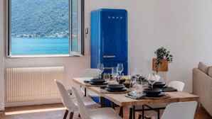 Uma área de refeições com uma mesa posta para quatro pessoas, uma geladeira azul, vista para um lago e montanhas, e uma planta no parapeito da janela.