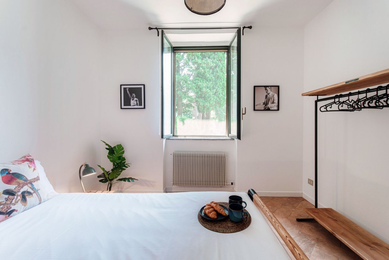 A bedroom with a bed, a window, a radiator, a plant, and a breakfast setup.