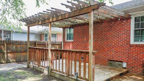 A backyard with a wooden deck, a pergola with string lights, a brick house, and a wooden fence. - Adorable pet-friendly fenced home in Hampton! Welcome to Redheart Cottage <3 (Hampton)