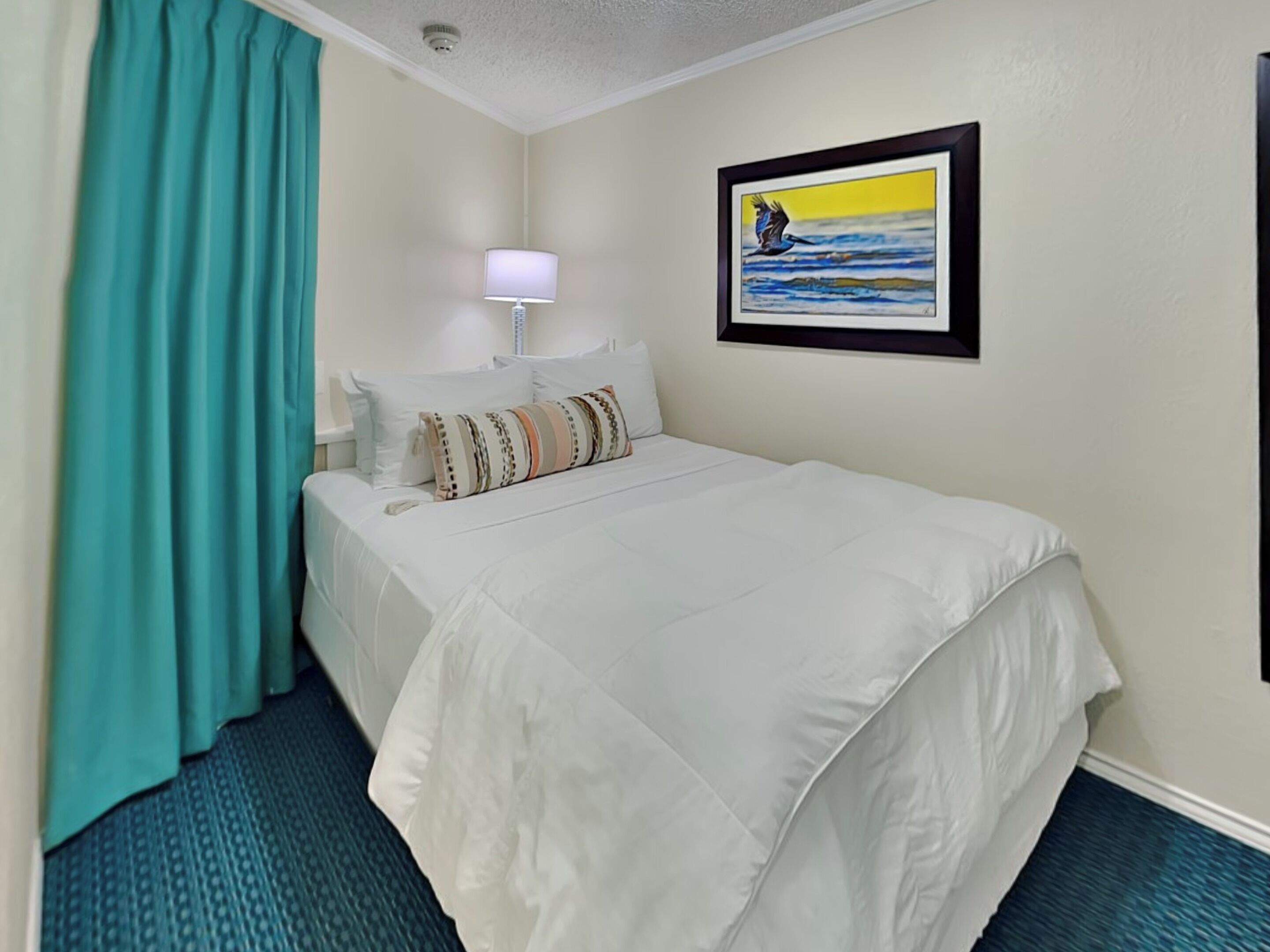 A hotel room with a bed, a nightstand, a lamp, a painting, and teal curtains.