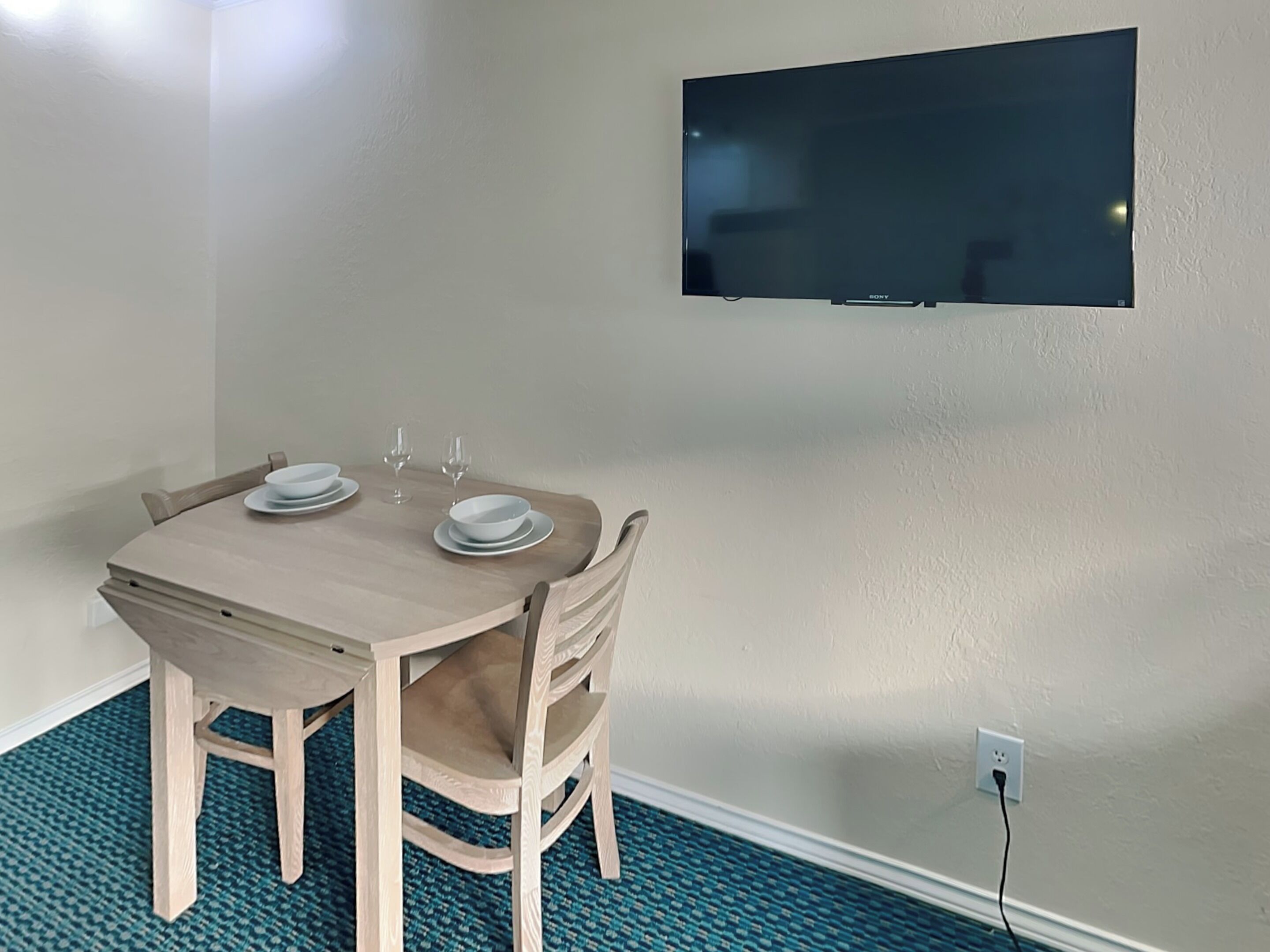 Een eethoek met een houten tafel gedekt voor twee personen, een wandtelevisie en een stopcontact in de muur.