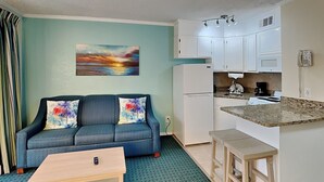 A compact living space with a sofa, a coffee table, a kitchen area with a refrigerator, stove, and microwave, and a painting on the wall. - Casa Del Mar 184 | Private Balcony, Partial Beach Views (Galveston)