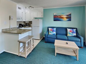 A compact living space with a kitchenette, a blue sofa with colorful cushions, and a small wooden table. - Casa Del Mar 211 | Private Balcony, Partial Gulf Views (Galveston)