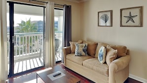 A living room with a beige sofa, a wooden coffee table, and a balcony with a view of palm trees. - Casa Del Mar 374 | Partial Gulf Views, Pool Access (Galveston)