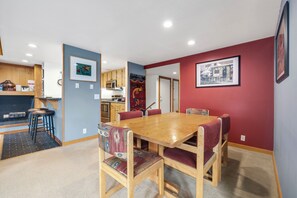 A dining area with a wooden table and chairs, a kitchen in the background, and a red accent wall. - Spacious 4-Bedroom in Keystone-Walk to lifts! (Keystone)