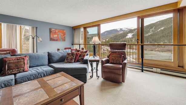 A spacious living room with a large window offering a mountain view, a blue sofa, a brown leather chair, and a wooden coffee table. - Spacious 4-Bedroom in Keystone-Walk to lifts! (Keystone)