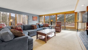 A spacious living room with a large sectional sofa, a coffee table, and a TV. The room has large windows offering a view of mountains. - Spacious 4-Bedroom in Keystone-Walk to lifts! (Keystone)
