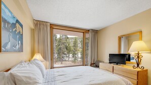 A hotel room with a large bed, a television on a wooden dresser, a painting on the wall, and a view of trees through the window. - Spacious 4-Bedroom in Keystone-Walk to lifts! (Keystone)