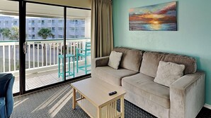 Ein Hotelzimmer mit Balkon, einem beigefarbenen Sofa, einem kleinen Holztisch und einem Gemälde von einem Sonnenuntergang.