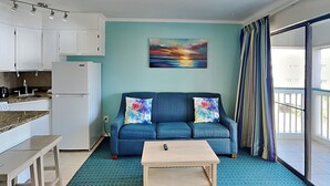 A compact living space with a kitchenette, a sofa with colorful cushions, a coffee table, and a large window with curtains. - Casa Del Mar 367 | Gulf Views, Pool Access (Galveston)