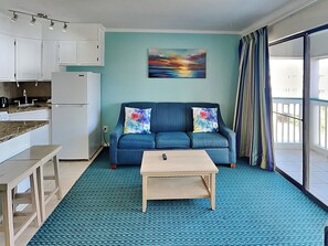 Ett kompakt vardagsrum med ett litet kök, en soffa med färgglada kuddar, ett soffbord och ett stort fönster med gardiner.