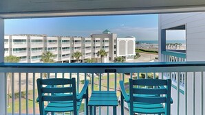 A balcony with two turquoise chairs and a view of a modern building complex with multiple balconies and palm trees. - Casa Del Mar 367 | Gulf Views, Pool Access (Galveston)