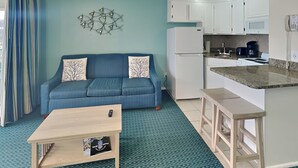 Een knus woongedeelte met een bank, een klein bijzettafeltje, een kitchenette met een koelkast, fornuis en magnetron, en een kamerbreed tapijt met een patroon.