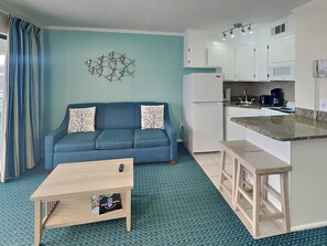 Ein kompakter Wohnbereich mit Sofa, kleinem Couchtisch, einer Küchenzeile mit Kühlschrank, Herd und Mikrowelle sowie einem gemusterten Teppich.
